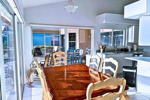 A dining area with a wooden table and chairs, a kitchen area with white cabinets, and a view through large windows. - Located Heart of the Island *Luxury Rebuild* Dock (Marco Island)