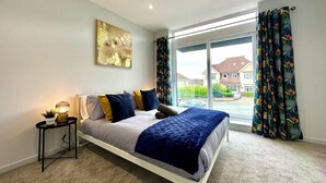 A bedroom with a bed, a bedside table, a lamp, a window with curtains, and a painting on the wall.