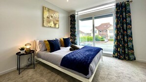 A bedroom with a bed, a bedside table, a lamp, a window with curtains, and a painting on the wall. - Stunning Brand New House - Sleeps 6 - Free Parking (Poole)