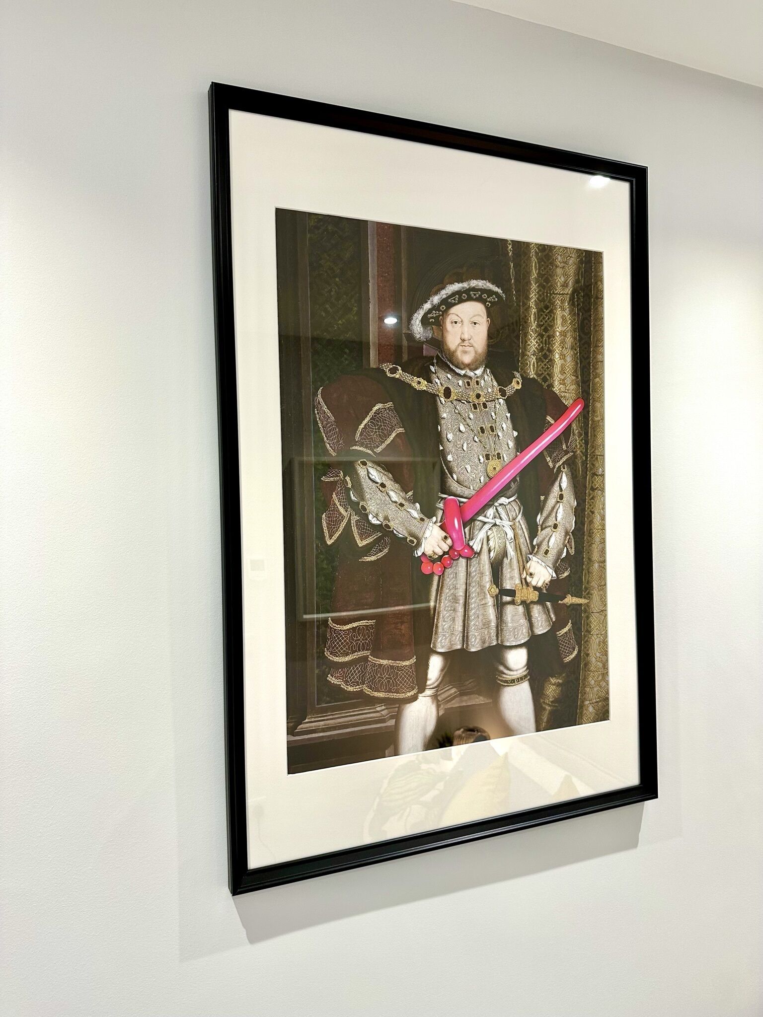 Un portrait encadré d’une figure historique, portant des vêtements somptueux, est accroché à un mur blanc.
