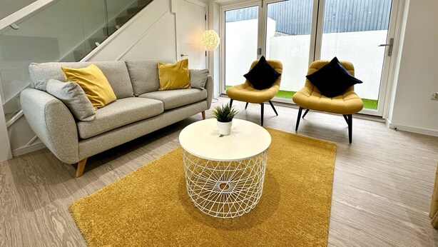 A modern living room with a grey sofa, two armchairs, a coffee table, and a staircase. - Stunning Brand New House - Sleeps 6 - Free Parking (Poole)