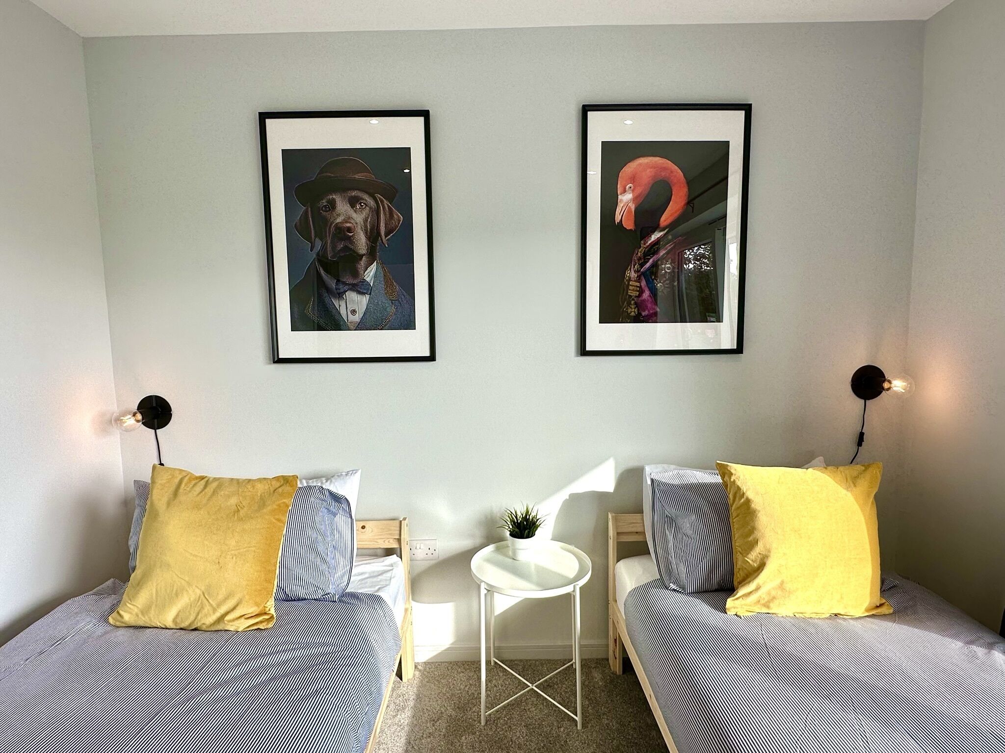 A room with two beds, each with a yellow pillow and a striped bedspread. There are two framed pictures on the wall, one of a dog and one of a flamingo. A small white table with a plant is between the beds.