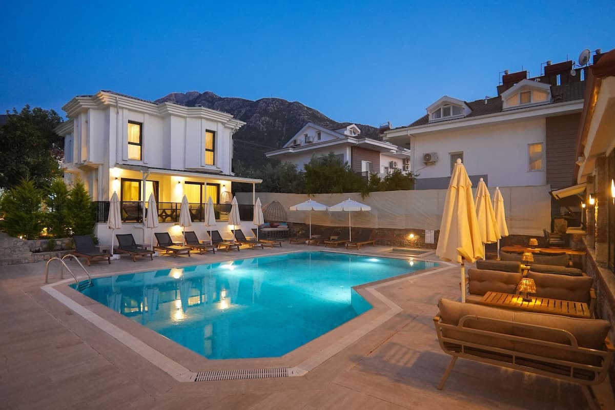A swimming pool with lounge chairs and umbrellas, a well-lit building, and mountains in the background.