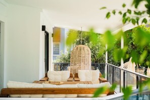 Een balkon met een houten bankje en witte kussens, een hangstoel van riet, en uitzicht op groen en een gebouw op de achtergrond.