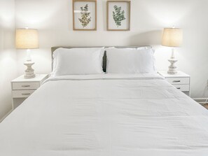 Un lit avec du linge de lit blanc, deux tables de chevet avec des lampes et deux gravures encadrées représentant des motifs botaniques accrochées au mur.