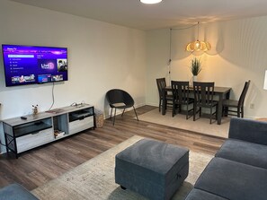 A modern living room with a flat-screen TV, a dining table with chairs, a sofa, and a small side table with a lamp.