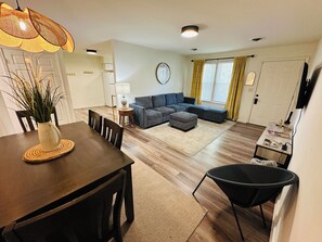 Een woonkamer met een bank, een poef en een eethoek met stoelen en een tafel.
