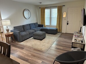 Un salon comprenant un canapé d’angle, une petite table ronde avec une lampe, un fauteuil en osier et une porte avec un porte-manteau.