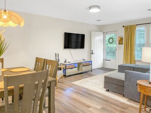 A modern living room with a dining area, a flat-screen TV mounted on the wall, and a door leading to an outdoor area.