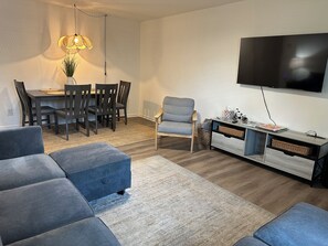 A modern living room with a dining area, a grey sofa, a TV mounted on the wall, and a wooden floor.
