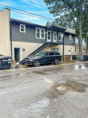 A two-story building with a staircase leading to the upper floor, a parked car, and a wet ground surface.