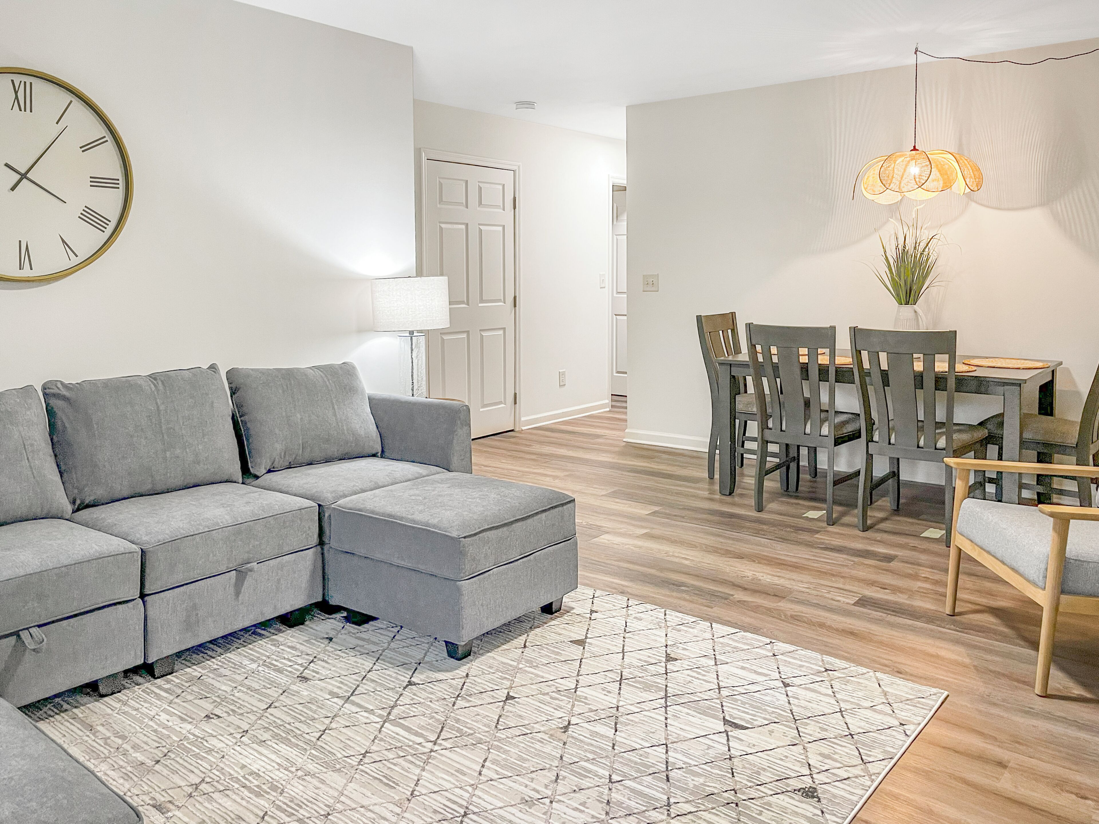 Un salon moderne avec un canapé d’angle, une table à manger avec des chaises, un tapis à motifs et une horloge murale.