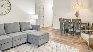 Ein modernes Wohnzimmer mit einem Ecksofa, einem Esstisch mit Stühlen, einem gemusterten Teppich und einer Wanduhr.