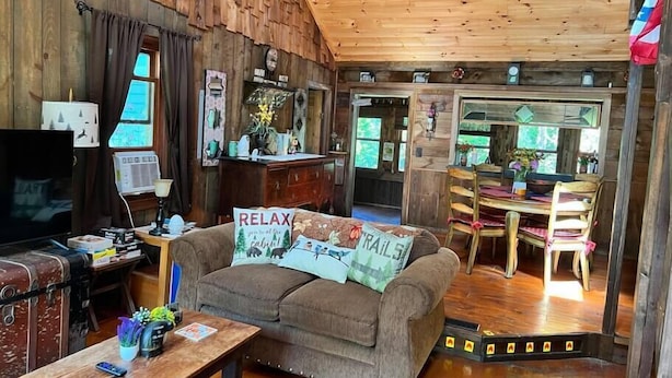 A cozy cabin interior with a wooden ceiling, a dining area with a table and chairs, a sofa with a 'RELAX' pillow, and a television on a wooden cabinet. - Cozy Cabin with Sauna & Firepit! (Monroe)