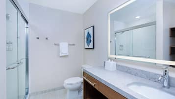 Un baño moderno con cabina de ducha de vidrio, inodoro, lavamanos con espejo grande y un cuadro en la pared.