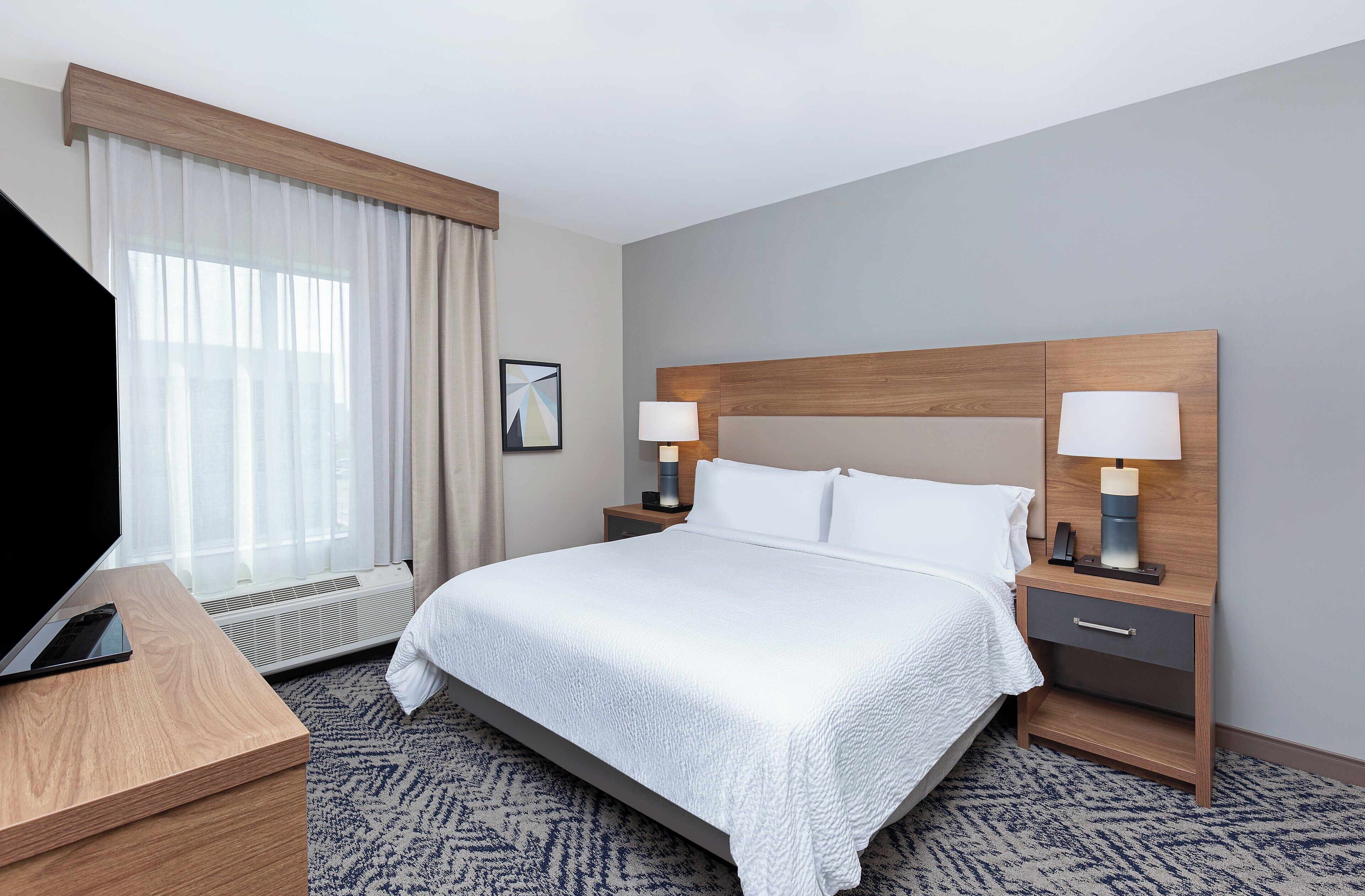 A hotel room with a large bed, two bedside tables with lamps, a flat-screen TV on a wooden desk, and a large window with sheer curtains.