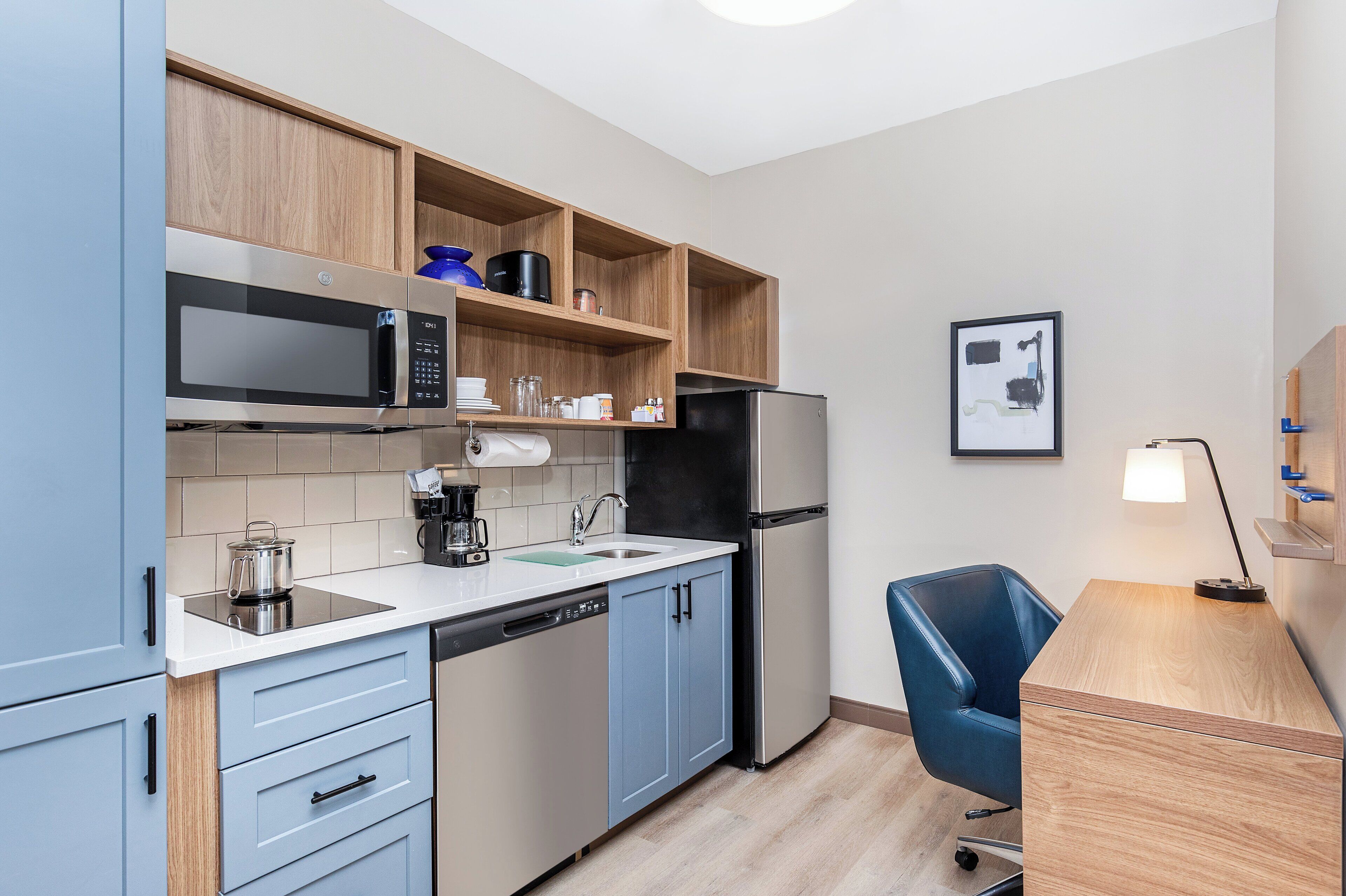 A compact kitchen with a microwave, stove, refrigerator, and a sink. There is a blue chair and a desk with a lamp.