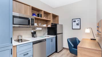 A compact kitchen with a microwave, stove, refrigerator, and a sink. There is a blue chair and a desk with a lamp.