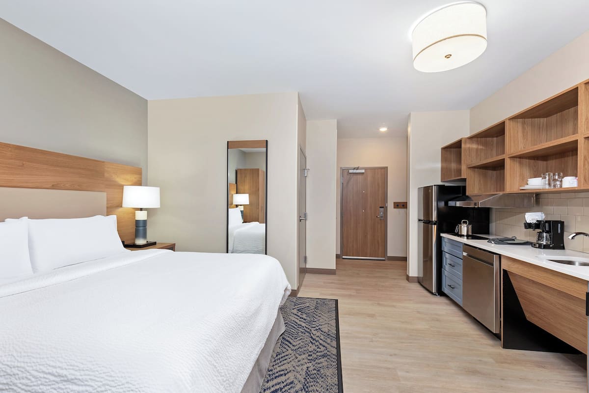A hotel room with a bed, bedside lamp, wooden headboard, a mirror, a door leading to a bathroom, a kitchenette with a stove, and a sink.