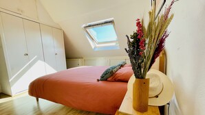 Quarto com teto inclinado, claraboia, cama com roupa de cama laranja, mesa de cabeceira de madeira e vaso com flores secas.