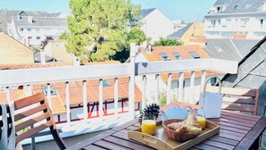 Mesa de madeira com bandeja de itens de café da manhã, incluindo croissants, suco e uma chaleira, em uma varanda com corrimão branco e vista para prédios e árvores.