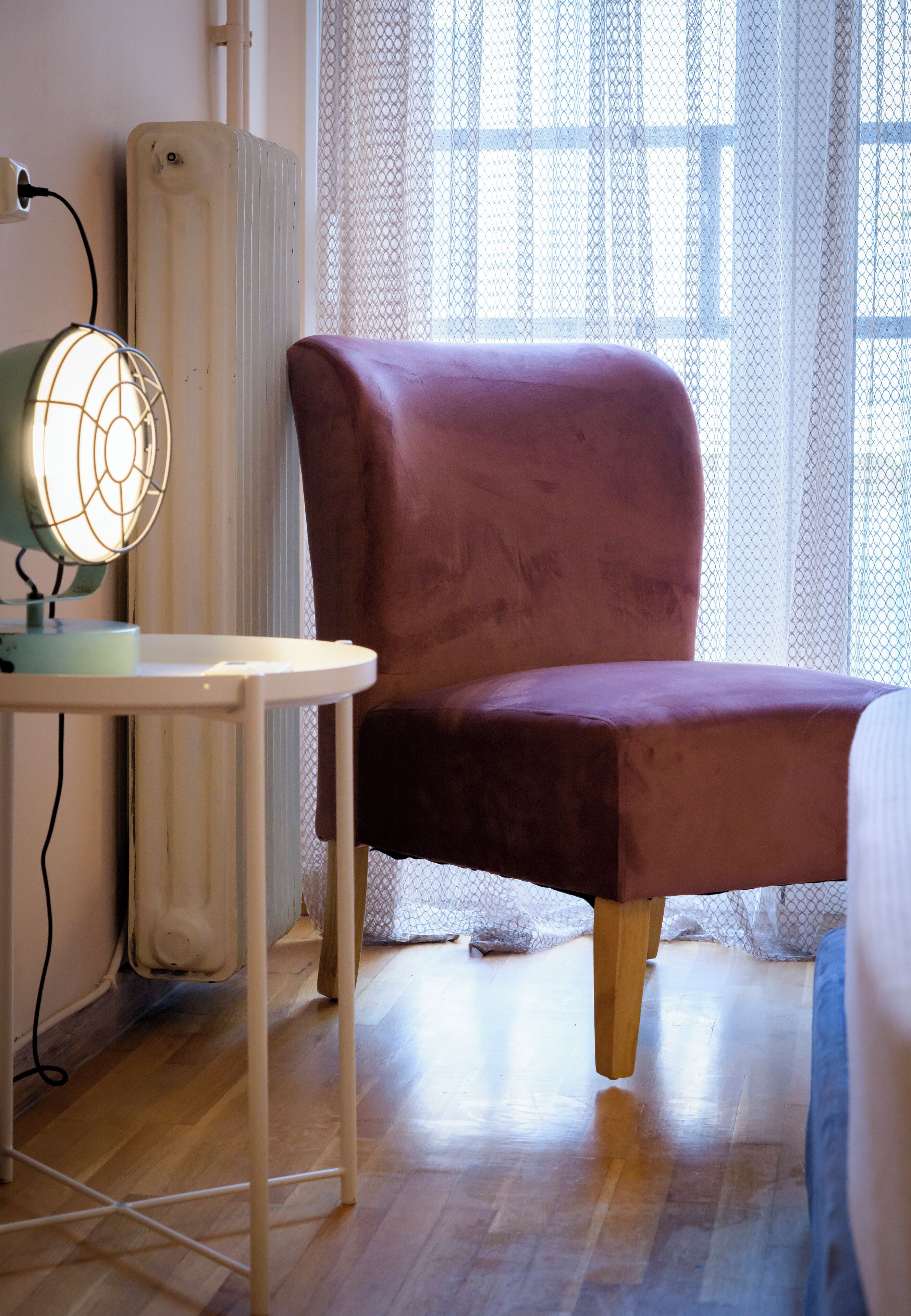 A room with a wooden floor, a radiator, a table with a lamp, and a chair.