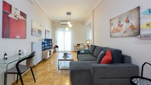 Ein modernes Wohnzimmer mit einem grauen Sofa, einem Glastisch und einem schwarzen Sessel. Ein Fernseher auf einem Schrank, eine Lampe und Kunstwerke an den Wänden sind ebenfalls zu sehen.