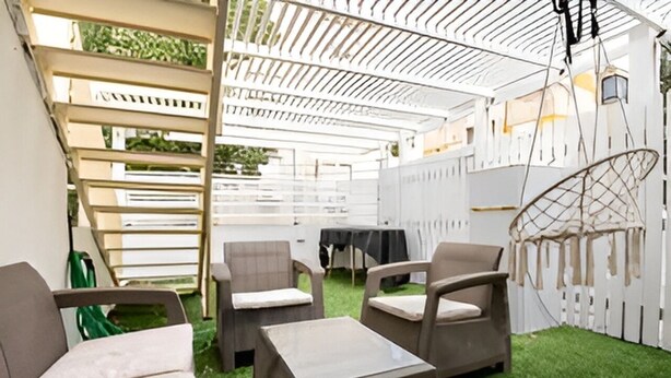 Een terras buiten met rieten meubilair, een witte pergola en een hangstoel.