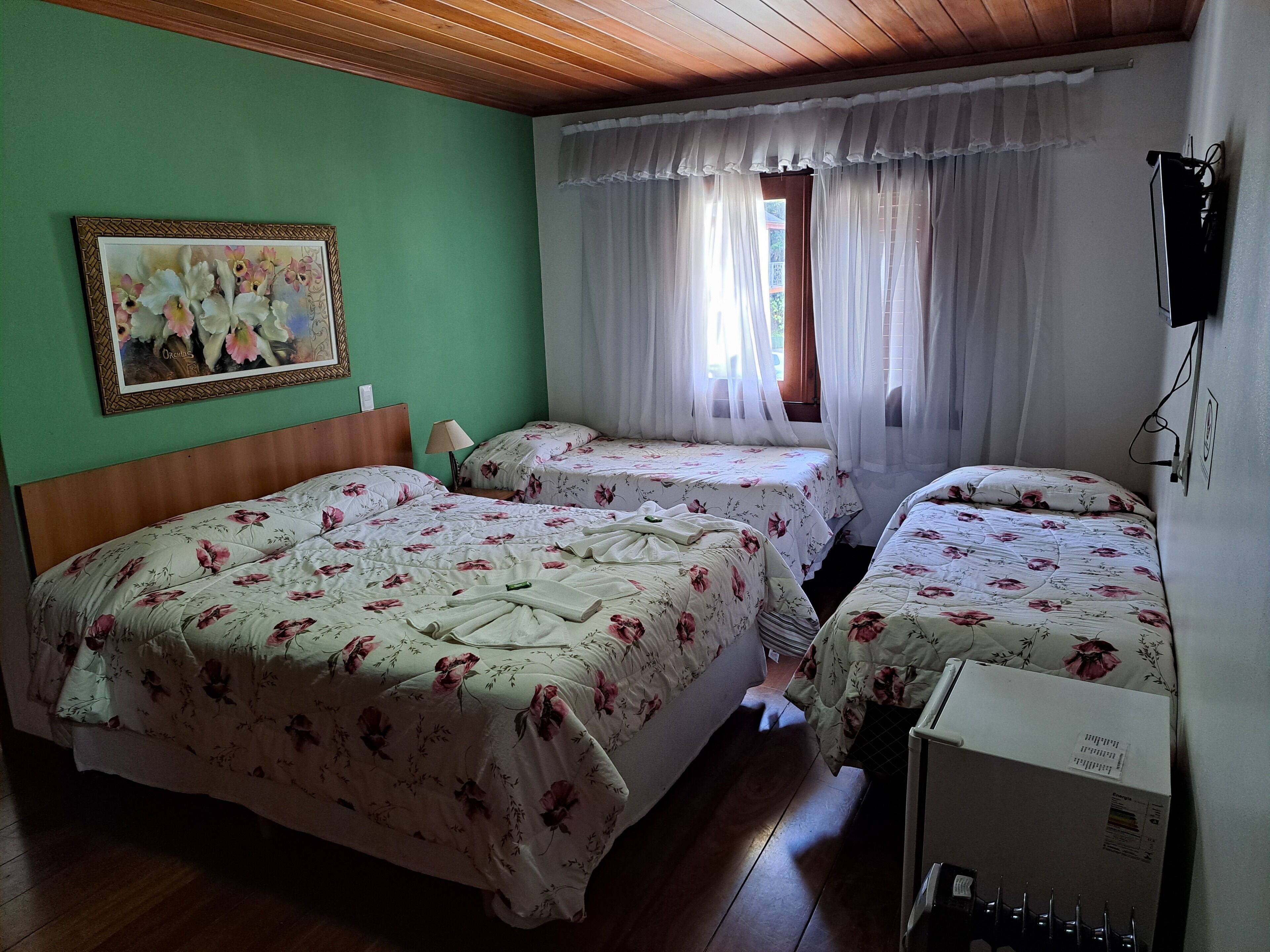 A room with two beds, a wooden ceiling, a green wall, a framed picture, a window with sheer curtains, and a TV mounted on the wall.
