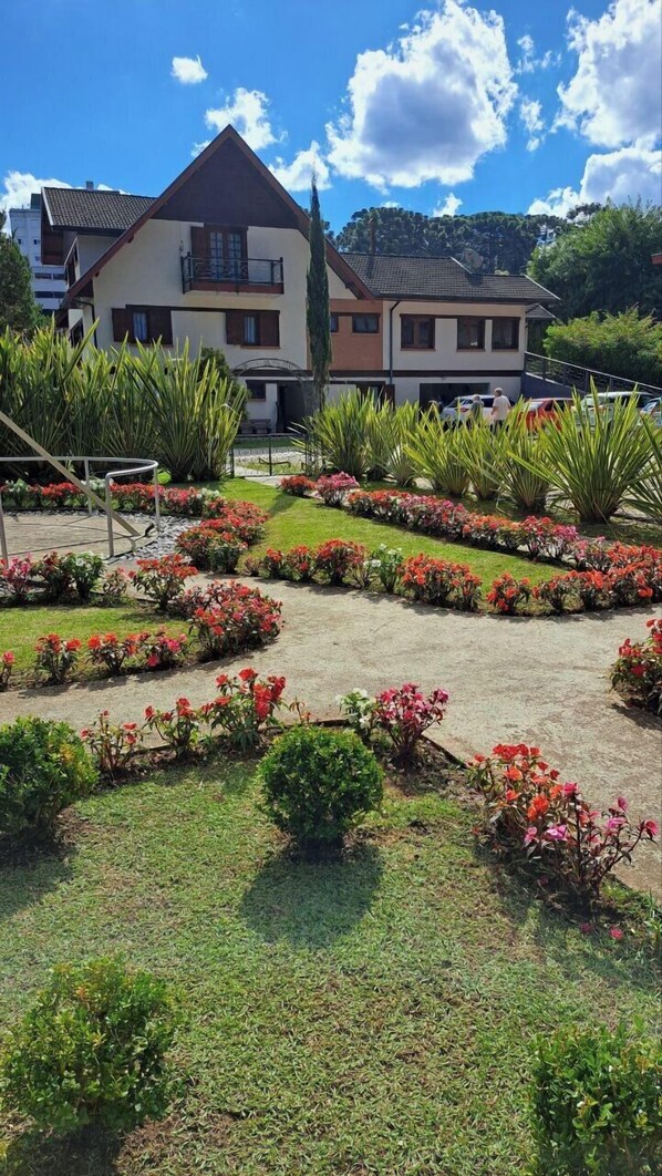 A residential area with a two-story house, a well-maintained garden with various plants and flowers, and a clear blue sky. - Pousada Recanto Feliz (Campos do Jordão)