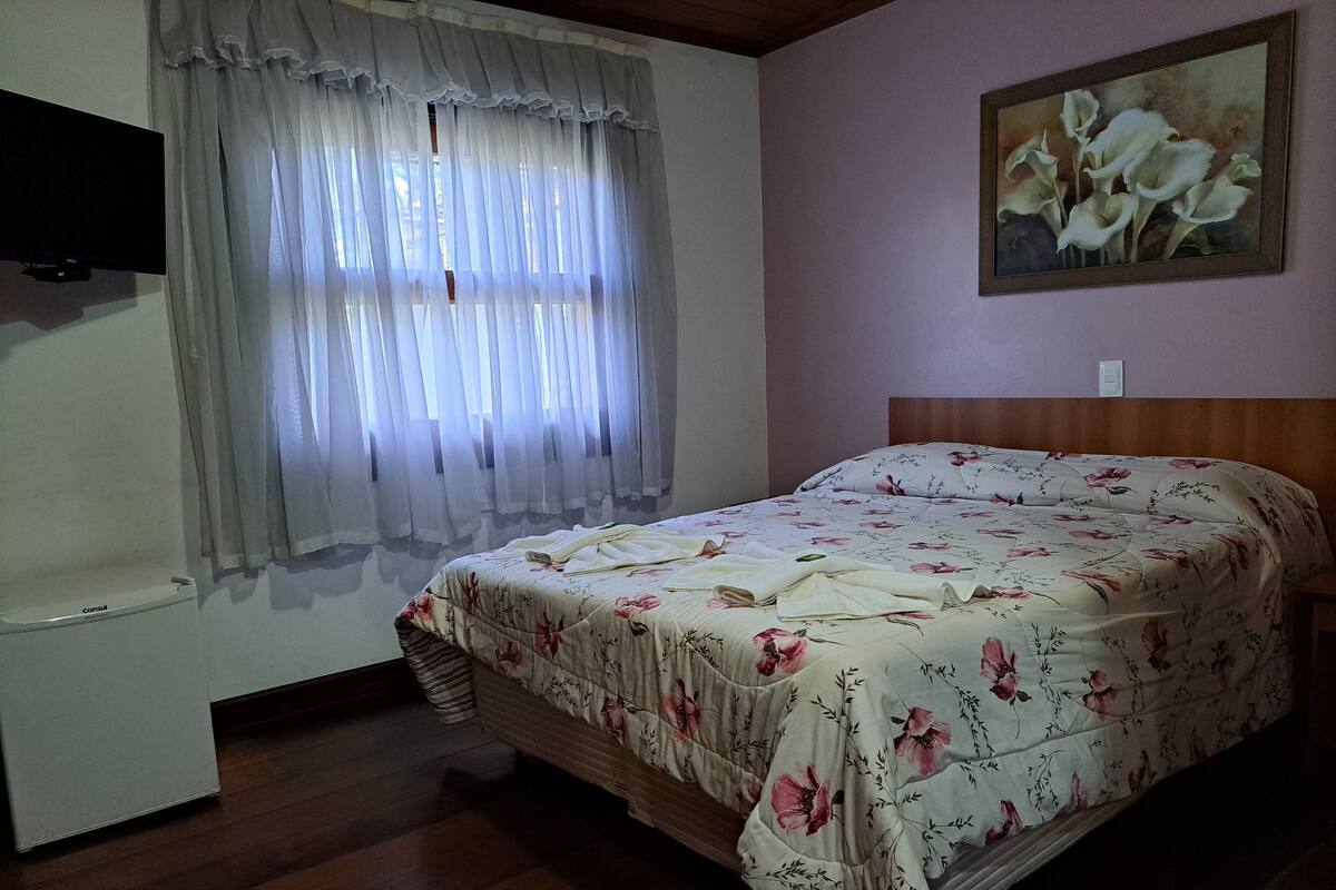 A bedroom with a bed, a painting, a TV, and a window with curtains.