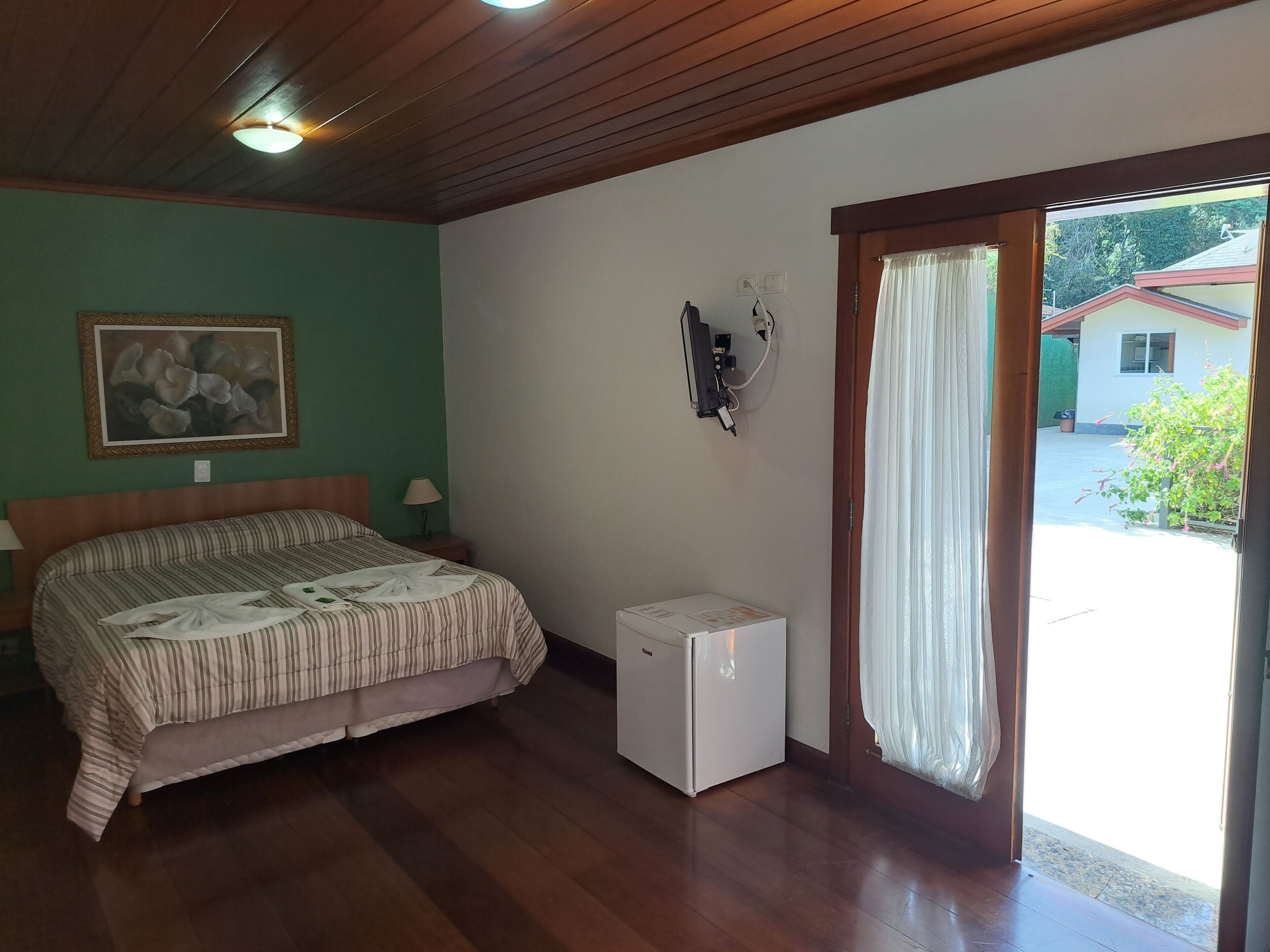 A hotel room with a bed, a small refrigerator, a painting on the wall, and a view of the outside.