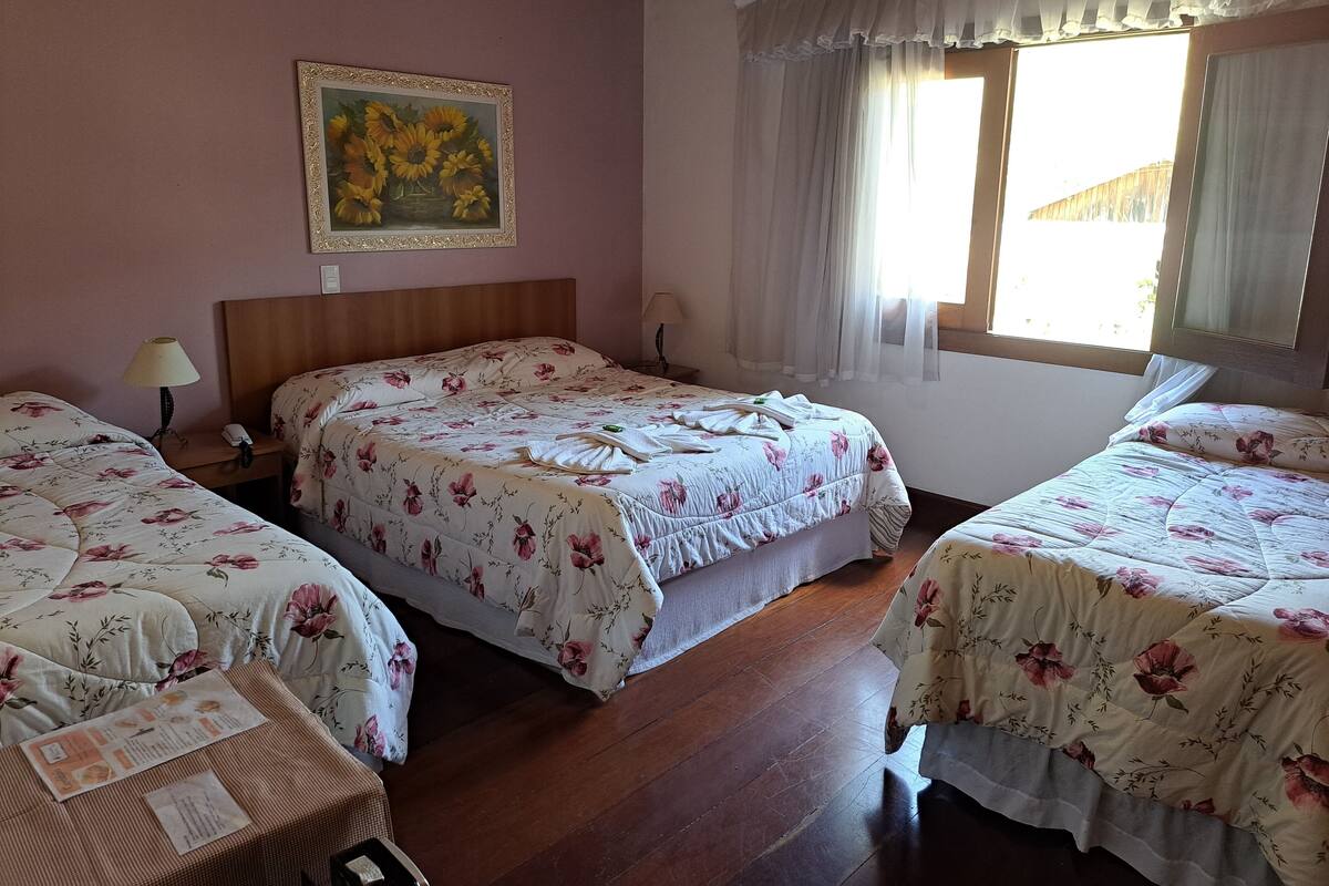 A hotel room with two beds, a wooden headboard, a painting of sunflowers, a window with curtains, and a bedside table with a lamp and a phone.