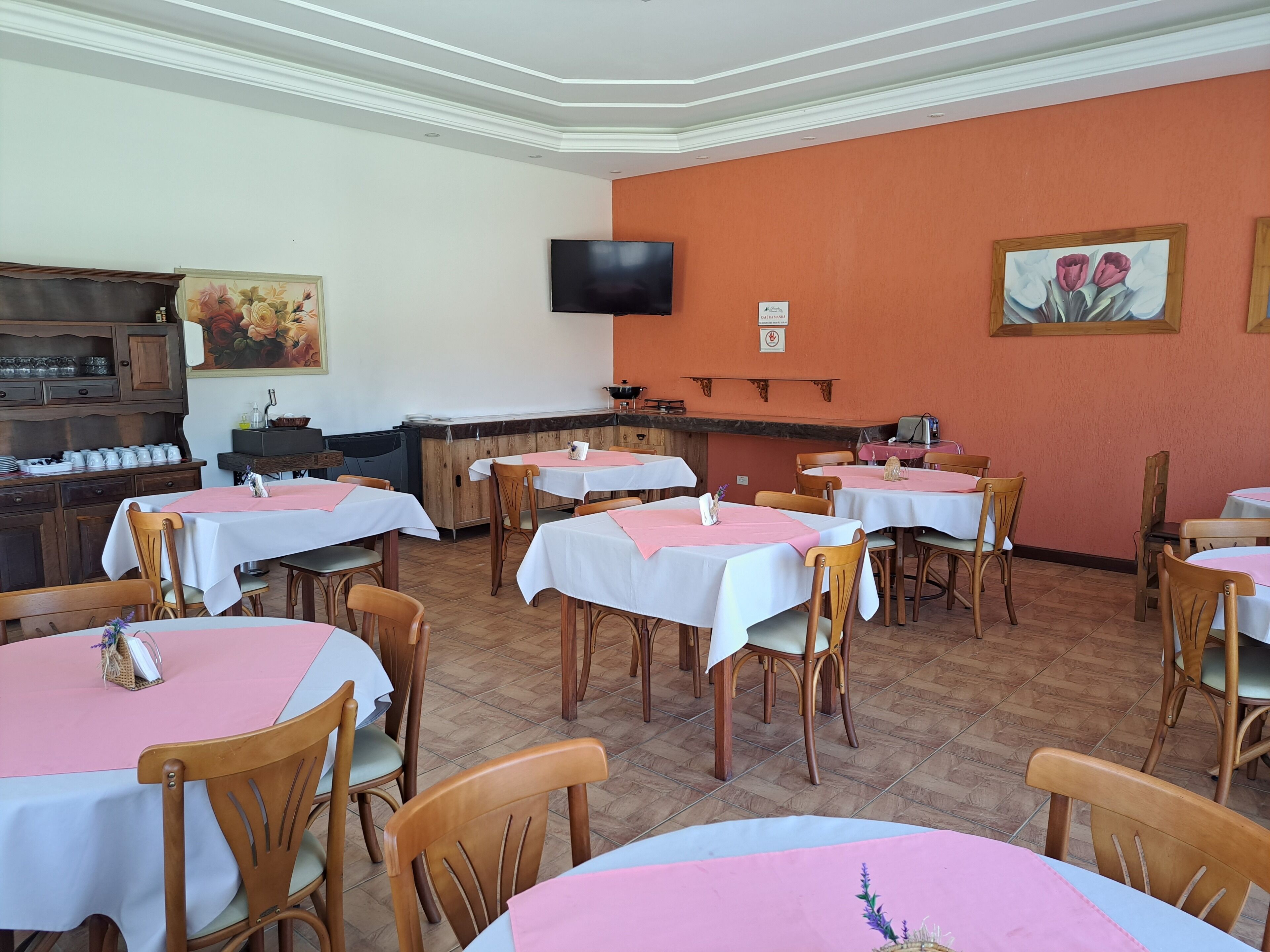 A restaurant with multiple tables and chairs, a television mounted on the wall, and a kitchen area with a wooden cabinet and a painting on the wall.