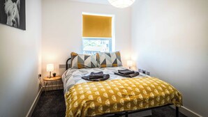 A neatly arranged hotel room with a bed, bedside lamps, a small table with a lamp, a window with a yellow blind, and a wall-mounted picture. - Two Bedroom Apartment, Sleep 5, Old Swan (Old Swan)