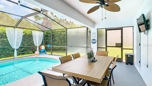 Un espace piscine intérieur moderne, doté d’une table et de chaises en bois, d’un ventilateur de plafond et d’une structure en verre fermée par des rideaux.