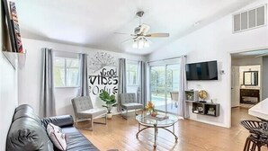 Un salon spacieux avec un canapé en cuir noir, deux fauteuils, une table basse et un ventilateur de plafond. Il y a une grande fenêtre avec des rideaux, un téléviseur à écran plat fixé au mur et un petit coin repas avec une table et des chaises.