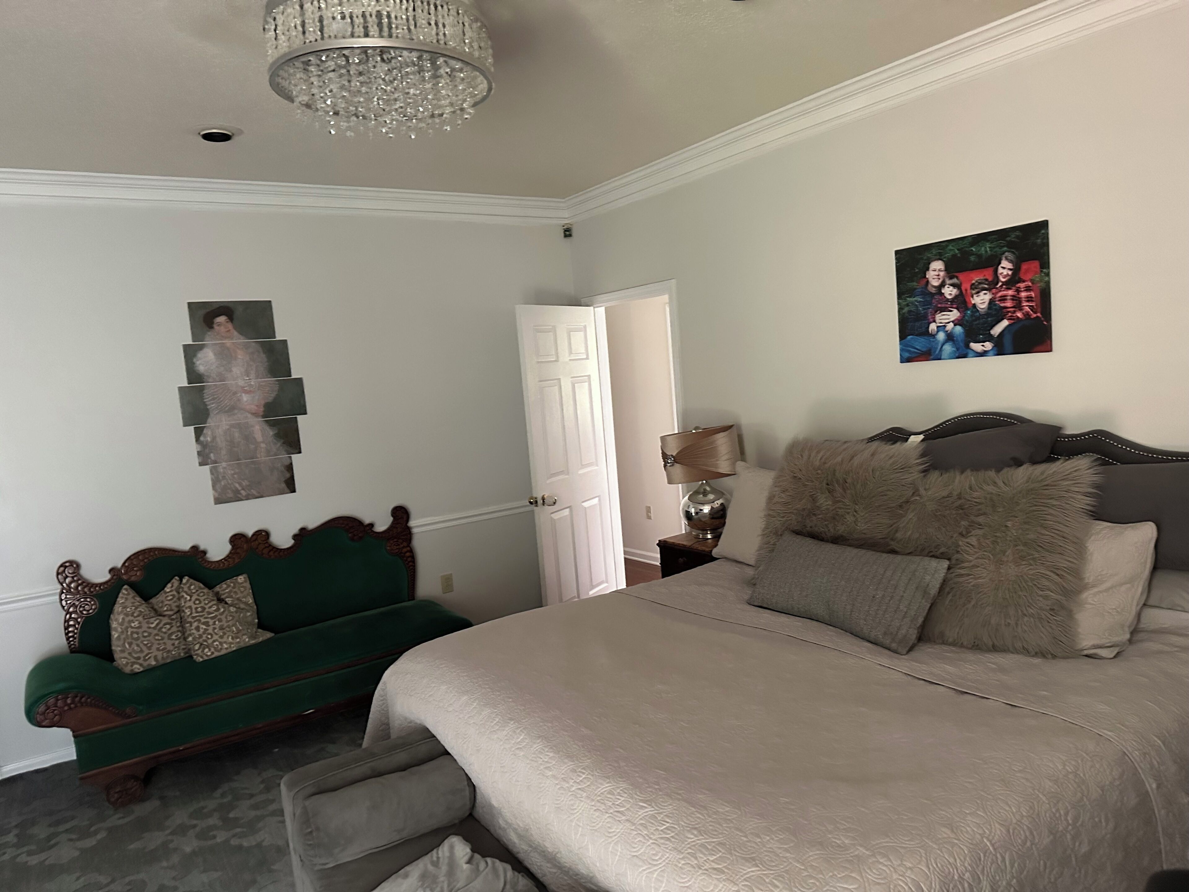 A bedroom with a large bed, a green sofa, a chandelier, a framed picture, and a lamp on a bedside table.