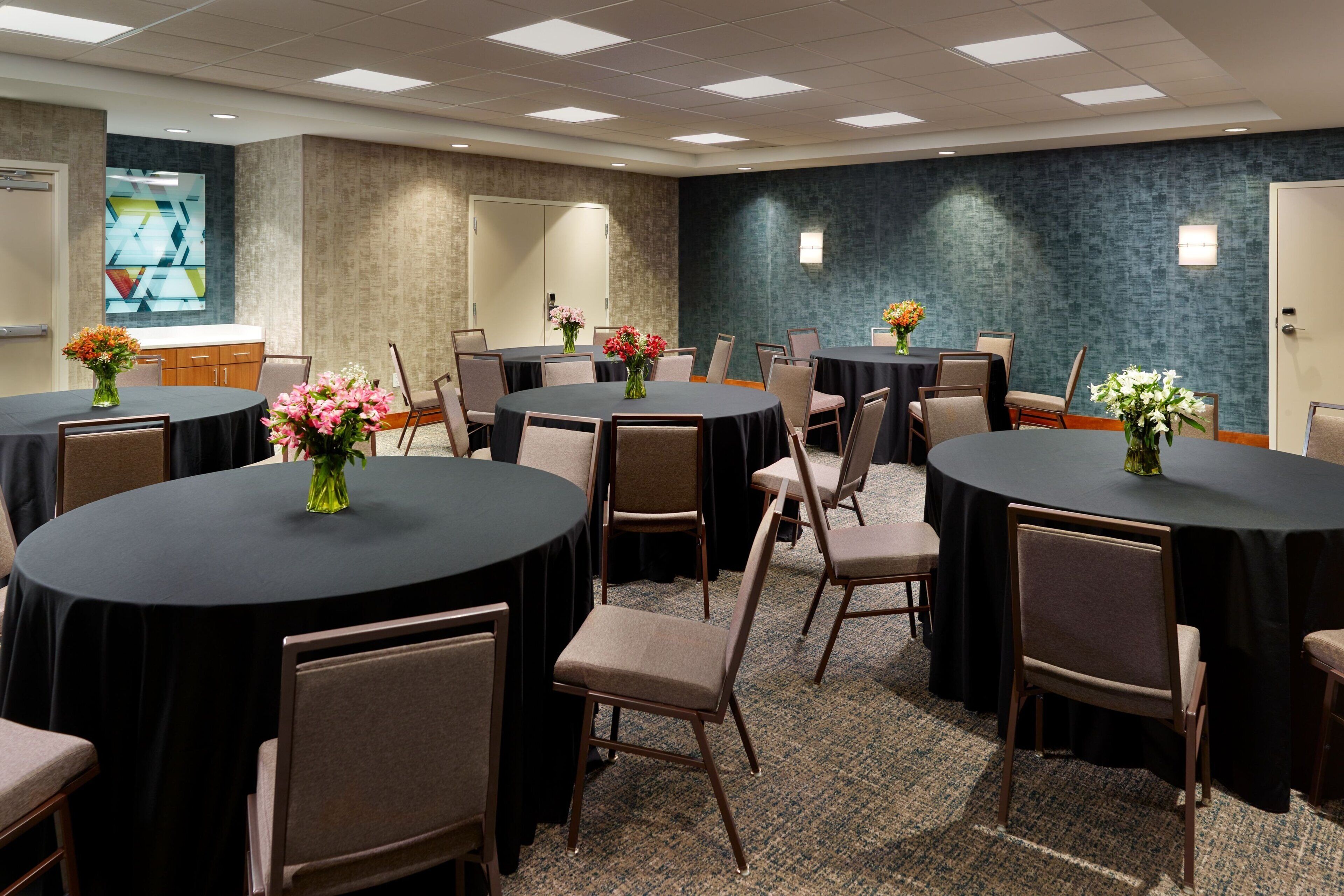 Un salón de conferencias con varias mesas y sillas, cada una con mantel negro y centros de mesa florales.