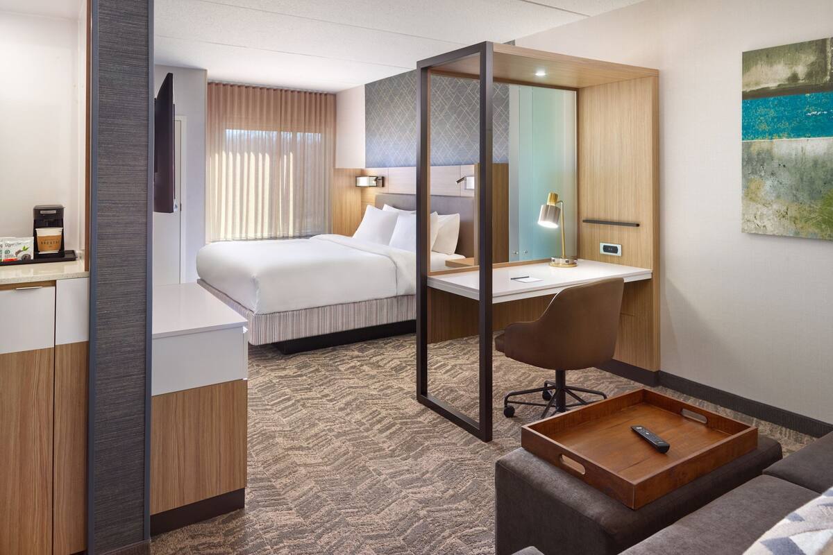 A hotel room with a bed, desk, chair, and a tray table with a remote control.