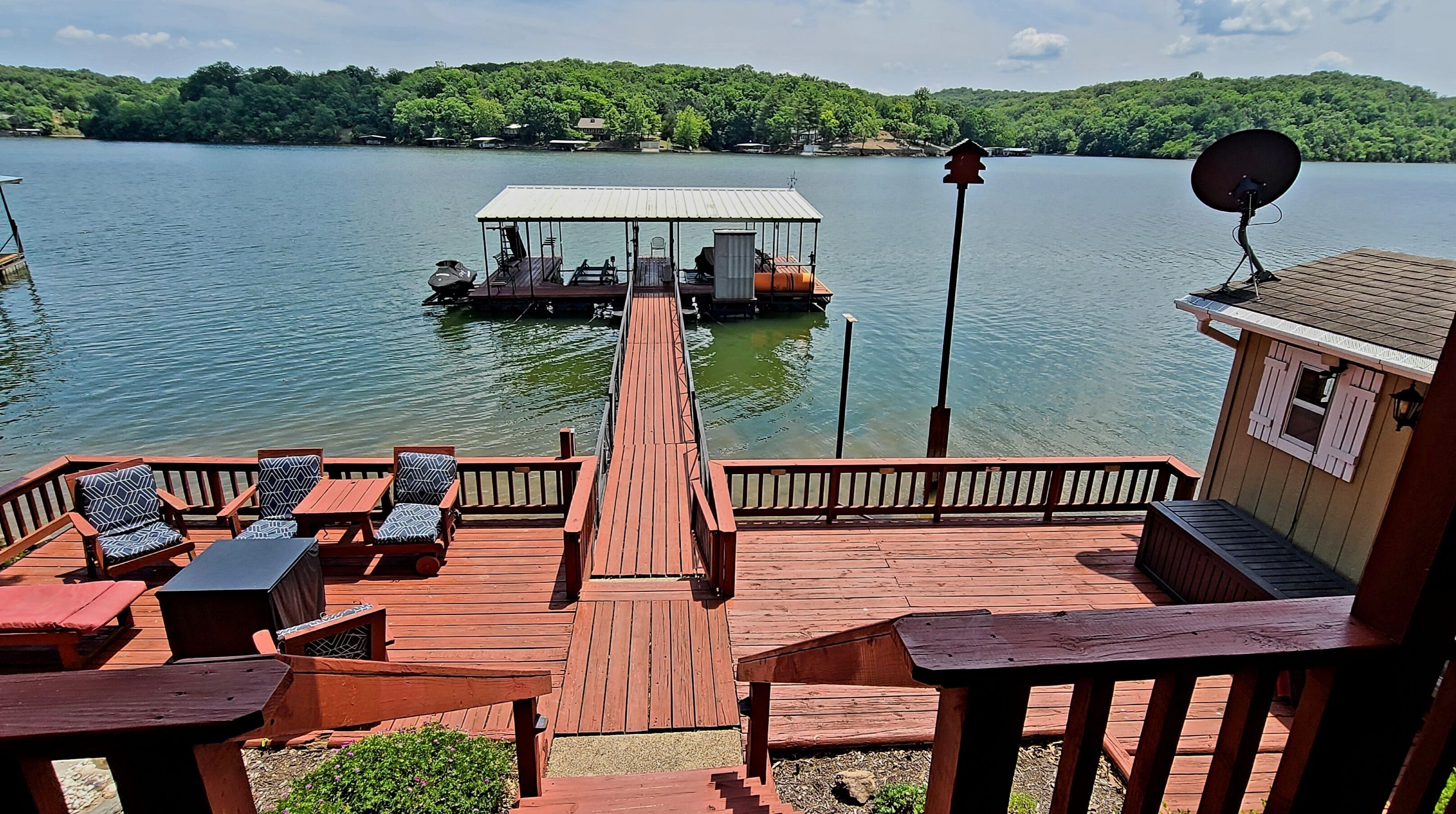 A wooden deck with outdoor seating, a dock extending into a lake, and a house with a satellite dish.
