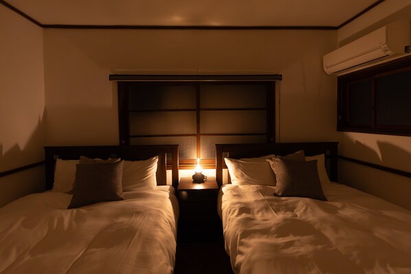 A hotel room with two beds, a nightstand with a lamp, a window, and an air conditioning unit. - Karakara (Osaka)