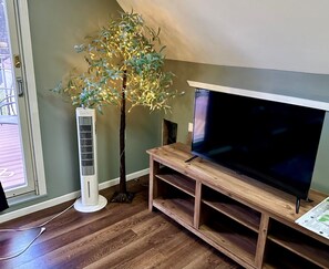 A living room with a large flat-screen TV on a wooden stand, a white standing fan, and a decorative tree with lights. - Modern Suite in Downtown Afton, 5min to skiing, hiking, riverboating & more! (Afton)