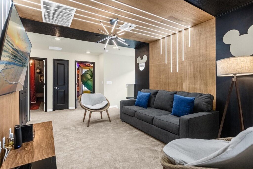 Un intérieur moderne avec un plafond en bois, un canapé gris agrémenté de coussins bleus, une chaise blanche et une décoration murale à l’effigie de Mickey Mouse.