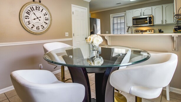 A dining area with a glass table surrounded by white chairs, a clock on the wall, and a kitchen in the background. - Soothing Savannah Townhome with Patio! (Savannah)