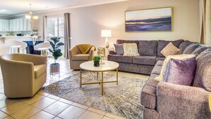 A living room with a sofa, armchair, and coffee table. There is a window with curtains, a ceiling fan, and a painting on the wall. - Soothing Savannah Townhome with Patio! (Savannah)