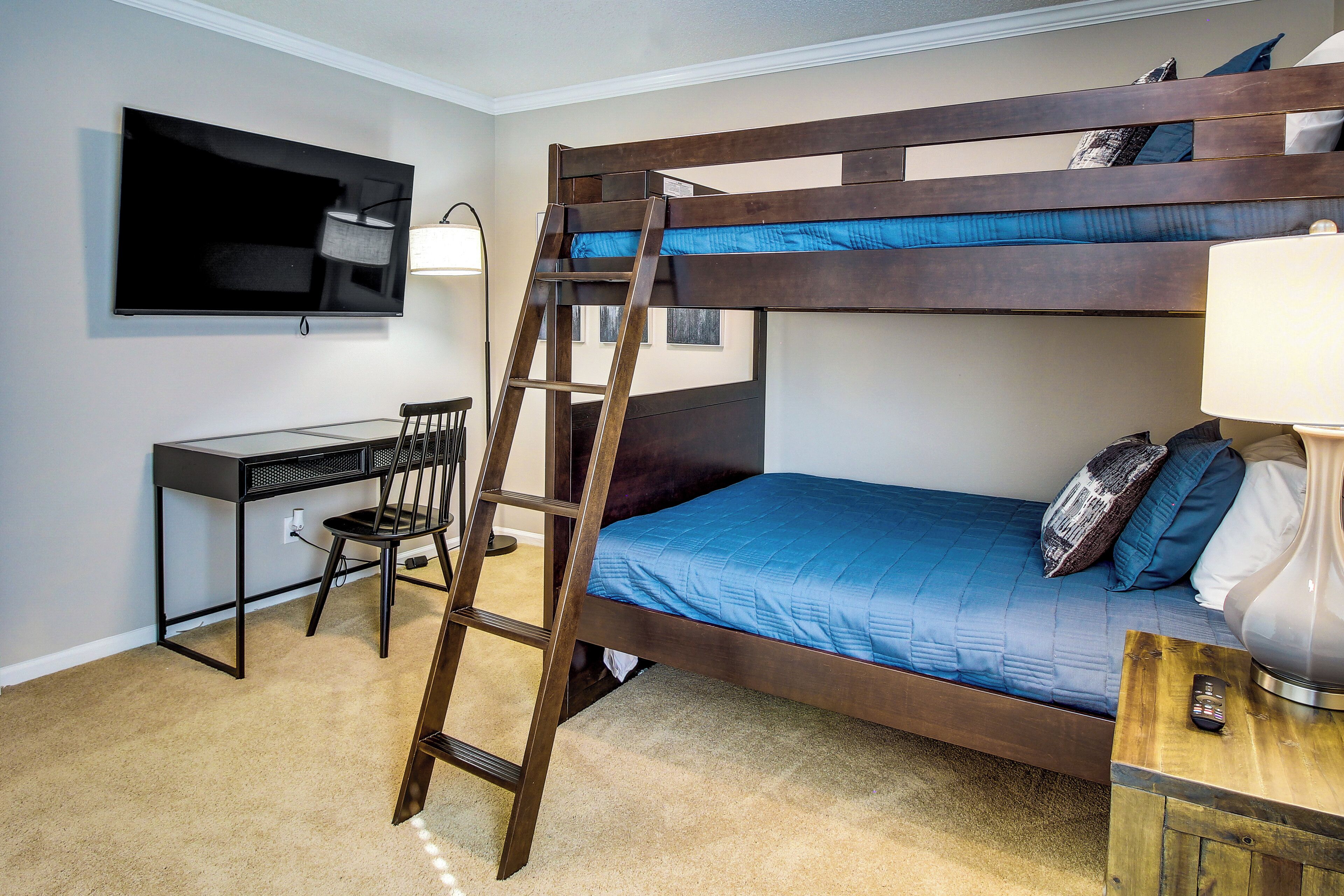 Un lit superposé avec un téléviseur fixé au mur, un bureau avec une chaise et une lampe sur une table de chevet.