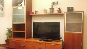 Ein Wohnzimmer mit einem Flachbildfernseher auf einer Holzkommode, einem Glastisch mit Pflanzen und einem geschlossenen Schrank.