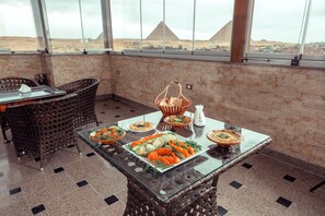 A dining area with a glass-top table set for a meal, wicker chairs, and a view of pyramids. - Brother Sphinx Inn (Giza)