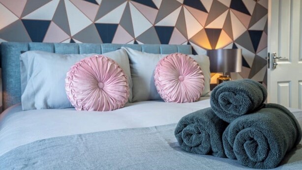 A neatly made bed with two pink pillows and a grey blanket, a blue headboard, and a geometric patterned wall. - Butterfly Gardens (Woodville)